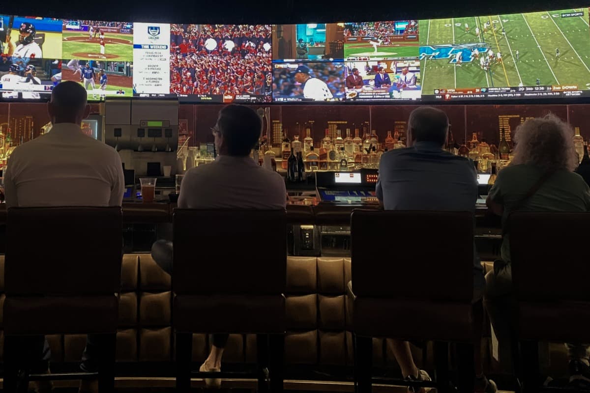 People sitting at a sports bar watching multiple live games on large screens above the bar.
