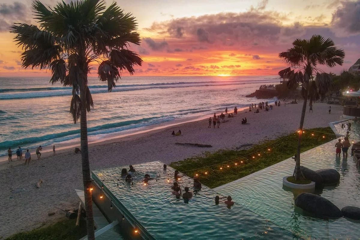 Beach club with infinity pool by the ocean at sunset, people relaxing while watching waves and sky.
