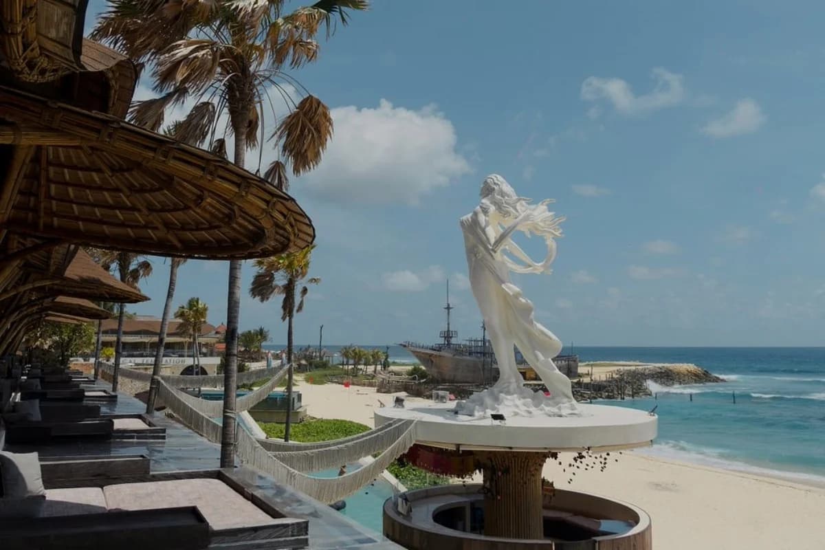 A Bali beach club overlooks a sandy beach and ocean, with shaded loungers, palm trees, and a large white statue.