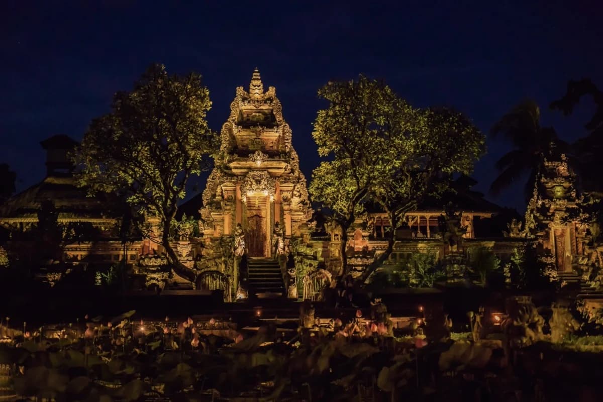 An ornate Balinese temple glows under night lighting, surrounded by trees and lotus-filled water in Ubud.