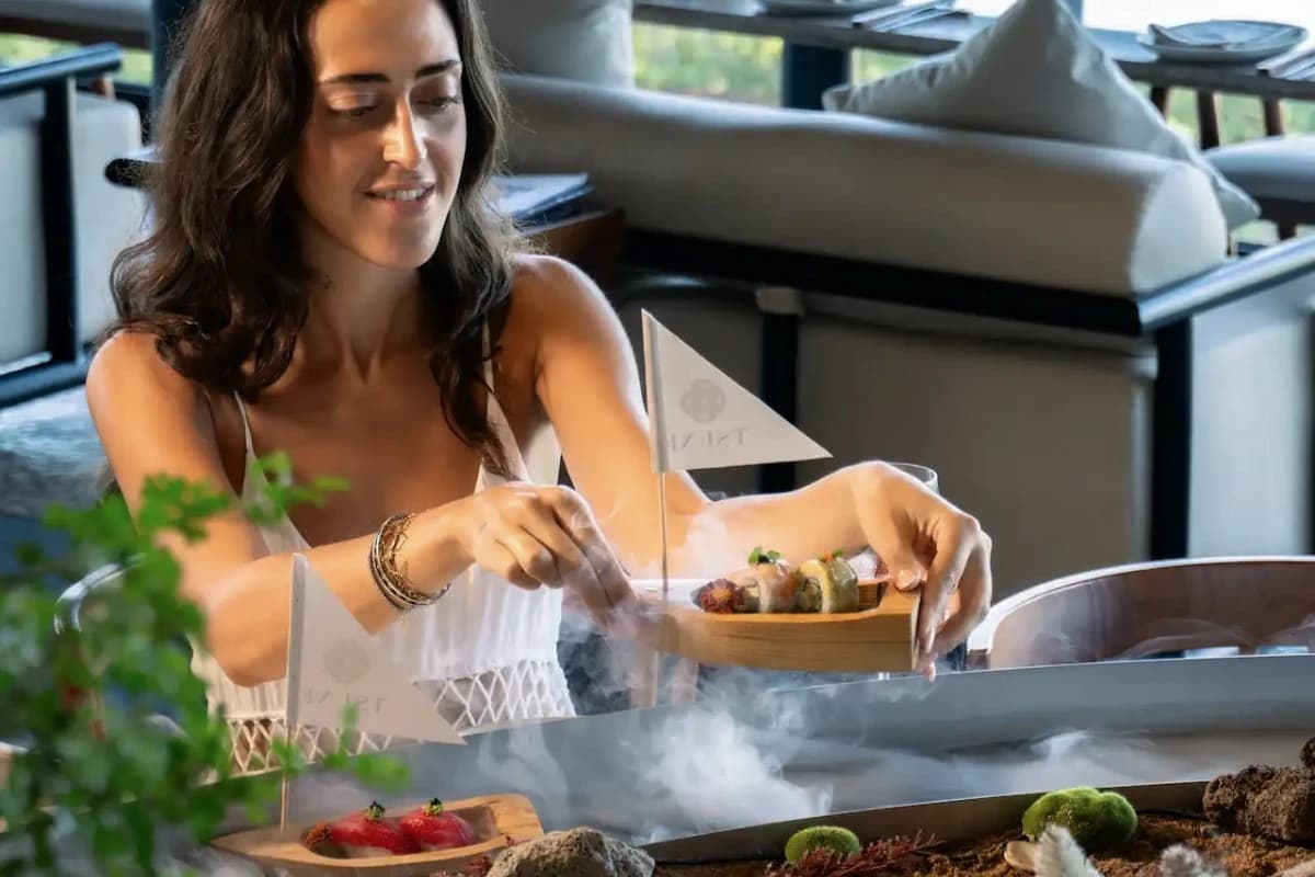 A woman serves sushi from a wooden boat on a water-filled table, illustrating the floating sushi dining experience at Tsune Sanur.