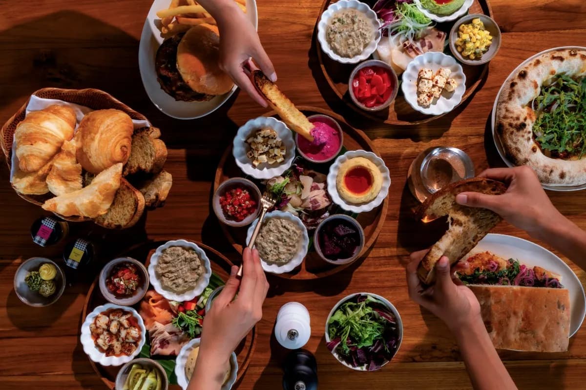 Hands share Italian-Mediterranean dishes, bread, salad, and sauces across a wooden dining table in Ubud.