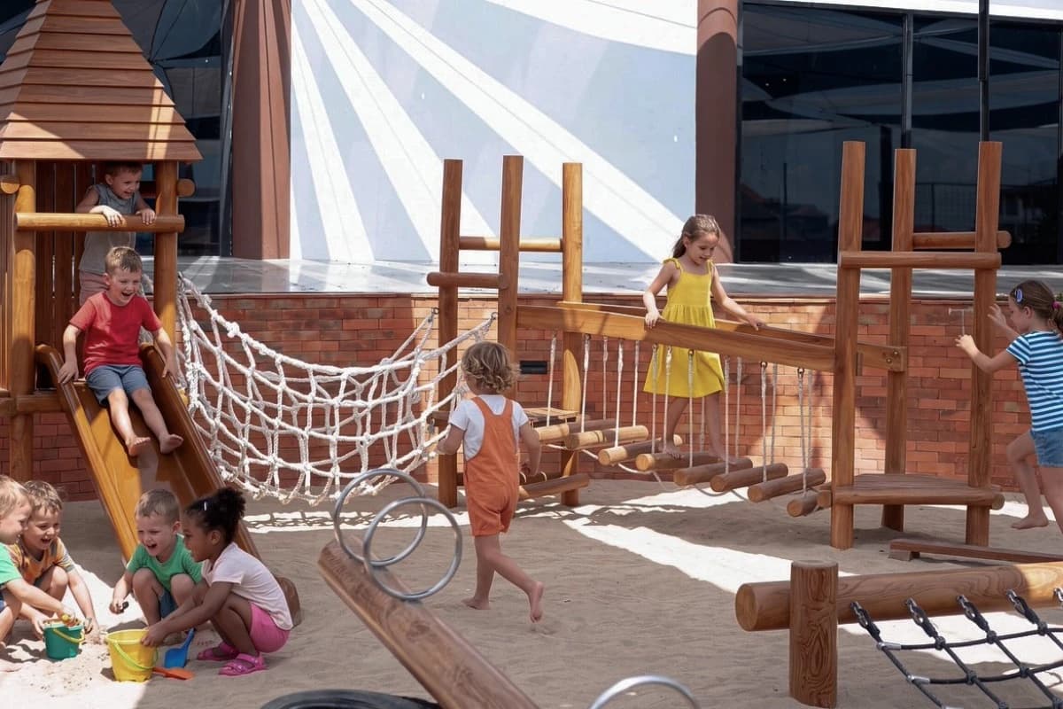 Children play on wooden slides, rope bridges, and climbing equipment in an outdoor playground with a sand area.