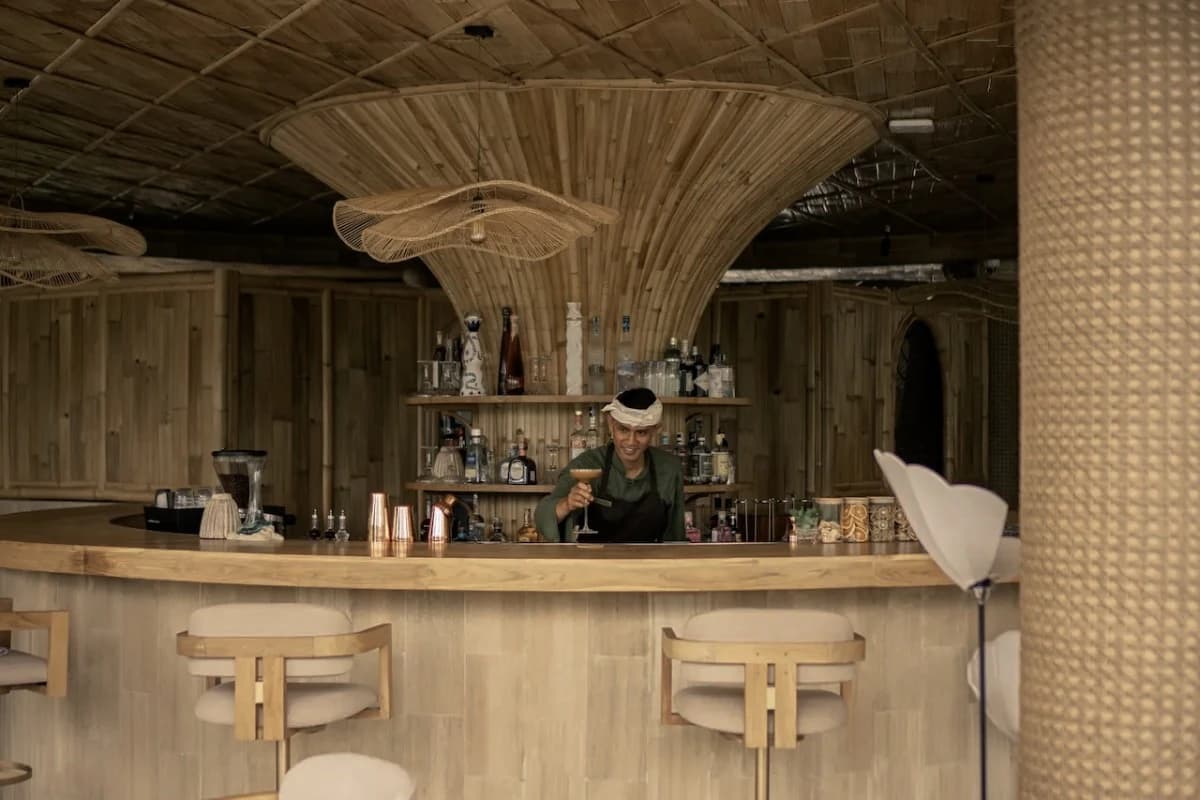 A bartender serves a drink at a bamboo bar with wooden stools, natural materials, and a warm interior in Ubud.