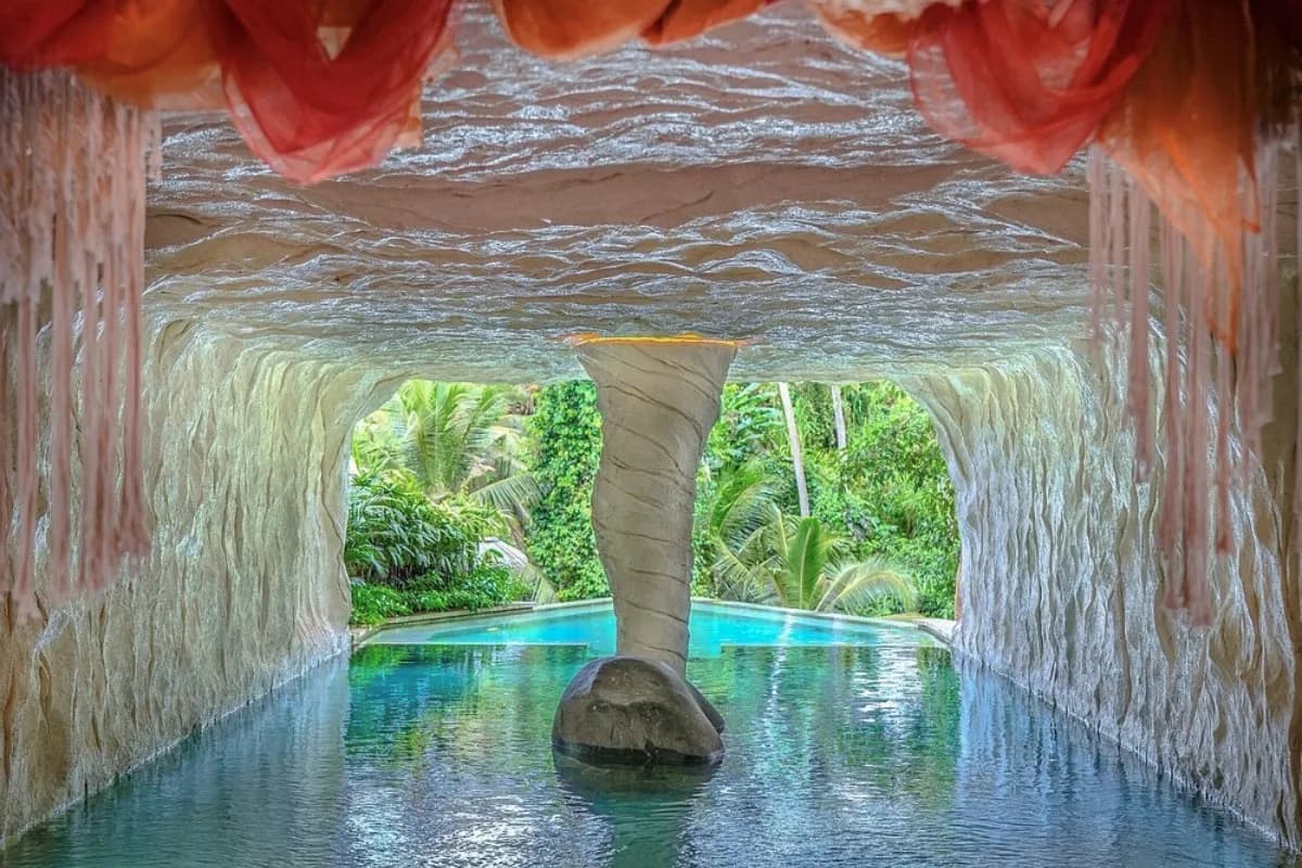 A turquoise swimming pool inside a rock cave opens onto lush jungle greenery and palm trees at a Ubud jungle club.
