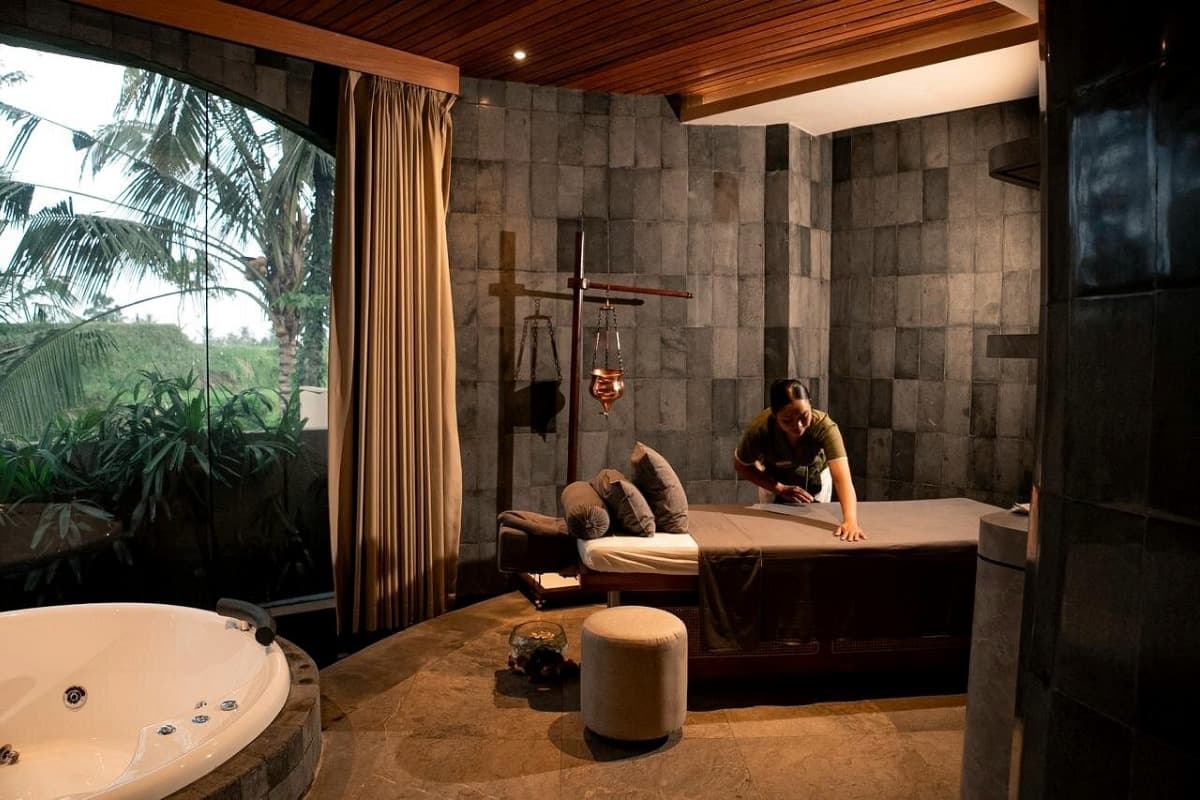 Spa therapist preparing a massage room in Bali with treatment bed, warm lighting, and garden view outside.