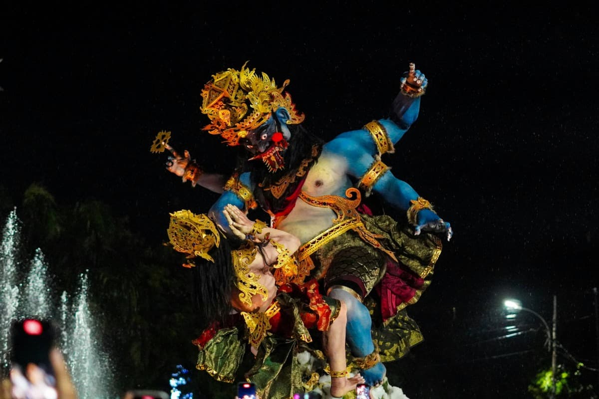 A giant, blue, multi-armed Ogoh-Ogoh demon statue crushes a smaller figure during Bali's Nyepi eve night parade.