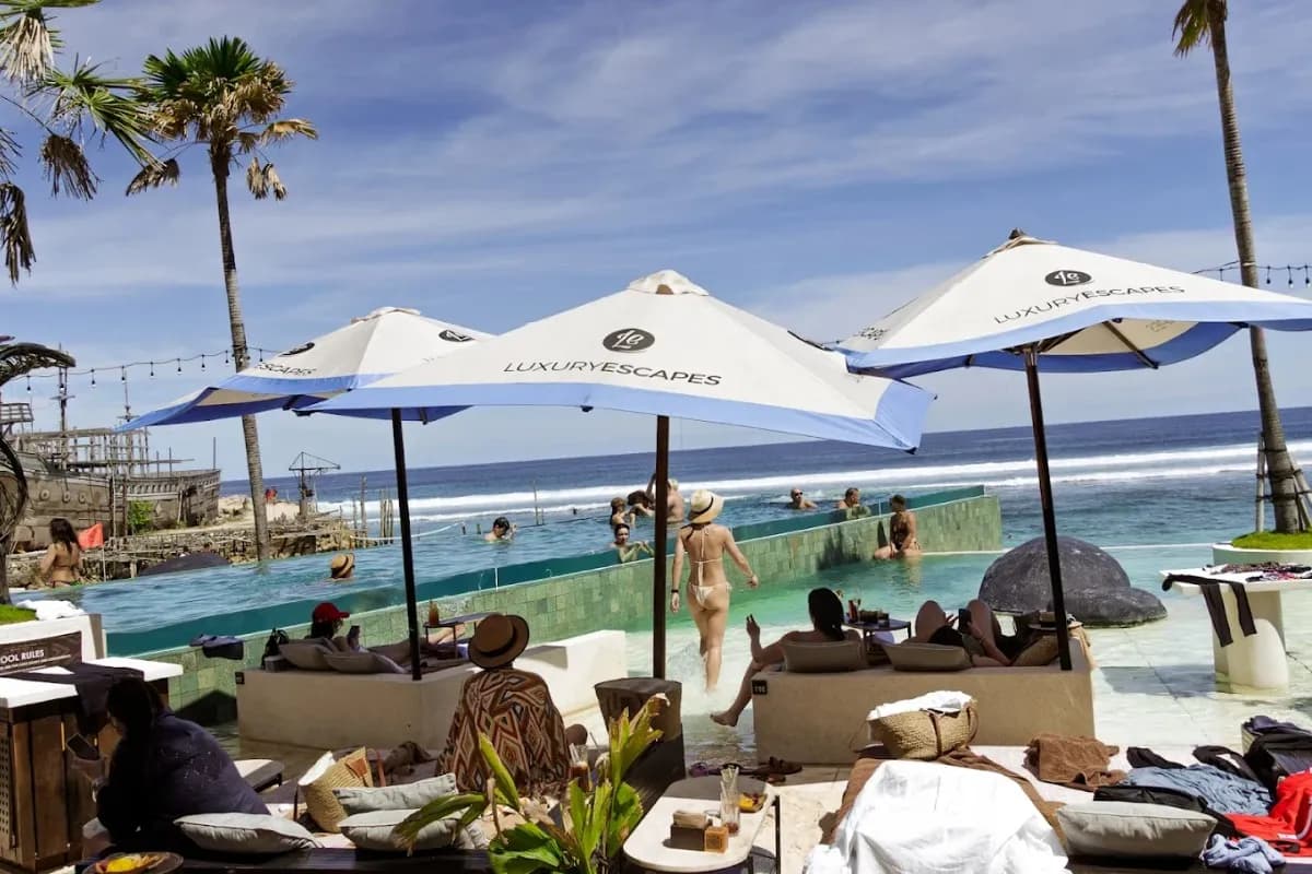 Beach club di Uluwatu dengan kolam infinity menghadap laut, pengunjung santai di bawah payung menikmati pemandangan.