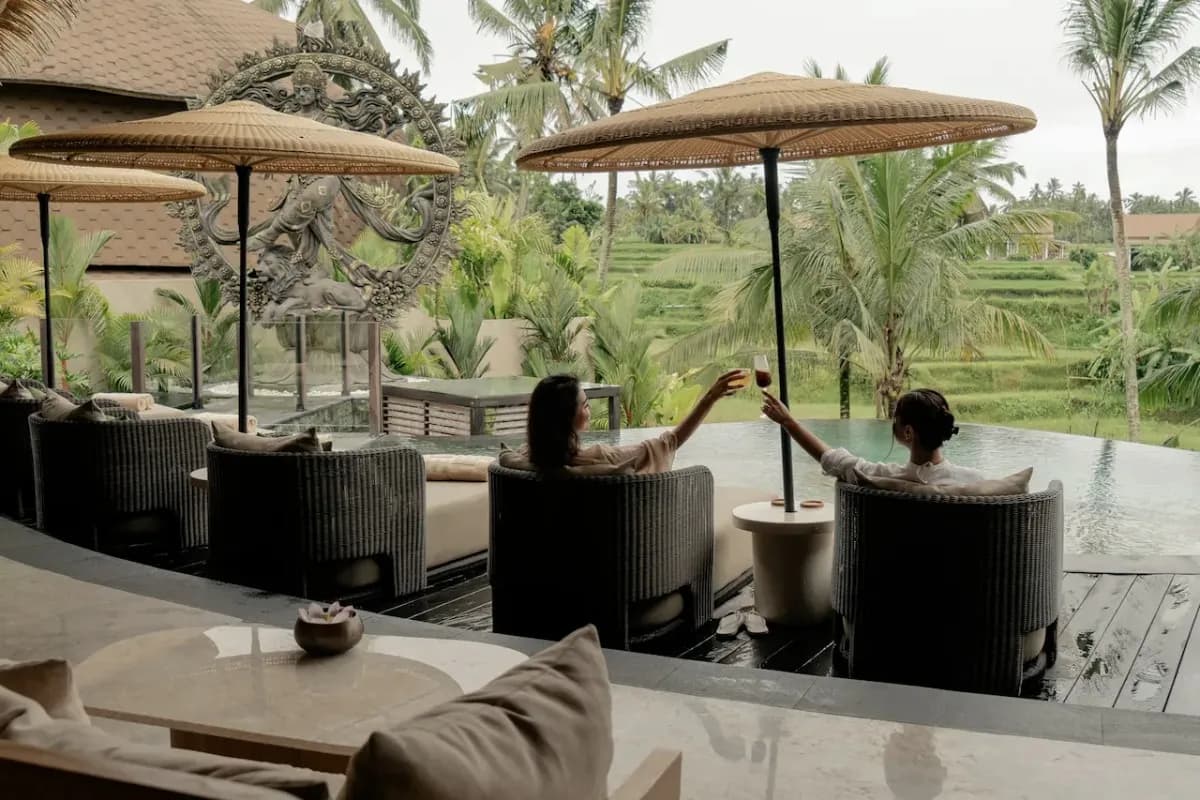 Two guests clink drinks beside an infinity pool overlooking Bali’s green rice fields and palm trees.