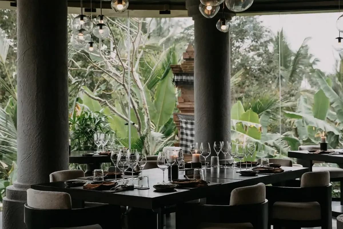 New Bali restaurant with elegant tables, wine glasses, pendant lights, and lush tropical greenery outside.