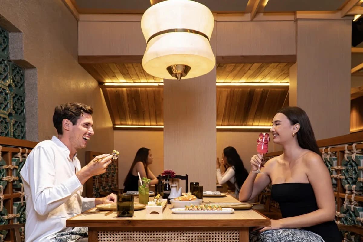 A couple enjoys Japanese dining at Shichirin Seminyak, sharing plated dishes and drinks at a wooden restaurant table.