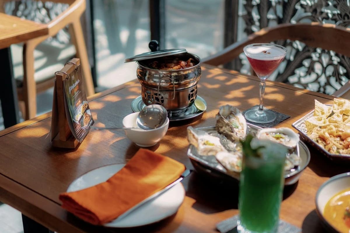 A wooden restaurant table displays Thai dishes, oysters, crispy snacks, and colorful drinks, illustrating a Thai dining experience in Sanur.